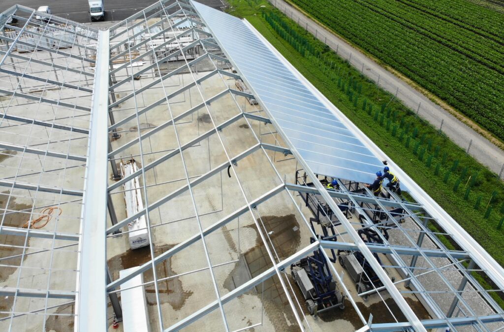 Montaż połaci dachowych przy użyciu podnośników, instalacja profili i elementów konstrukcji dachu nad częścią ogrodniczą.