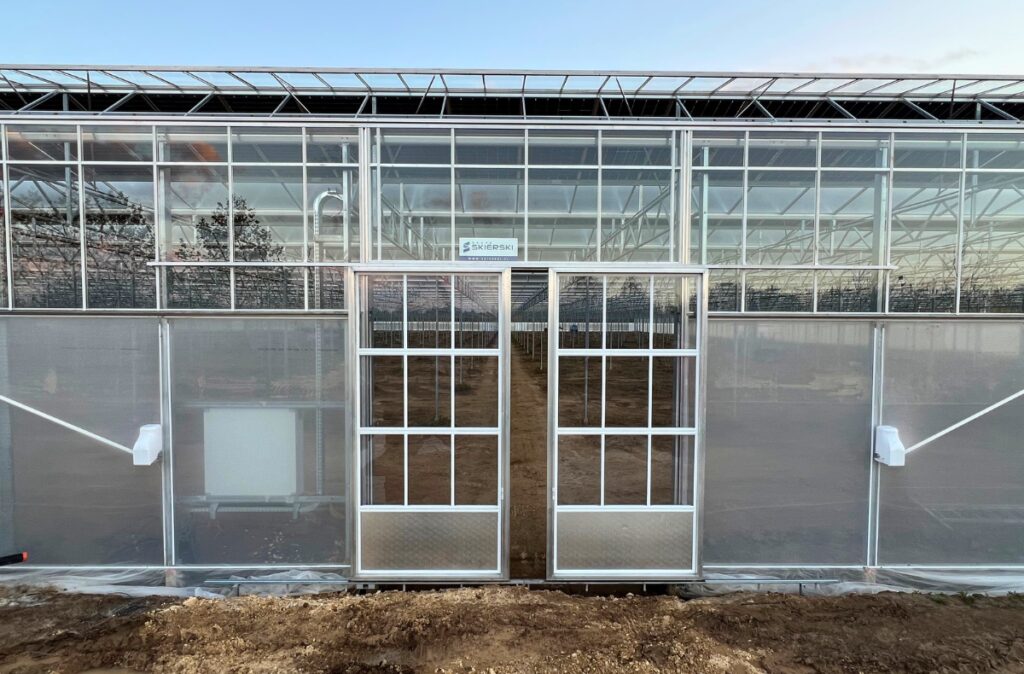 Wejście do szklarni po zakończonym szkleniu dachu, ścian i szczytów, z widocznymi drzwiami oraz elementami konstrukcyjnymi.