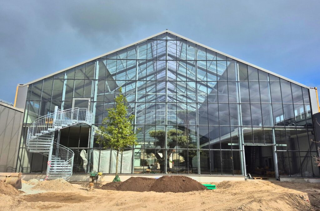 Szklana fasada wewnętrznej części ogrodniczej Ostmann, z widoczną dużą kubaturą i drzewem w centralnej strefie.