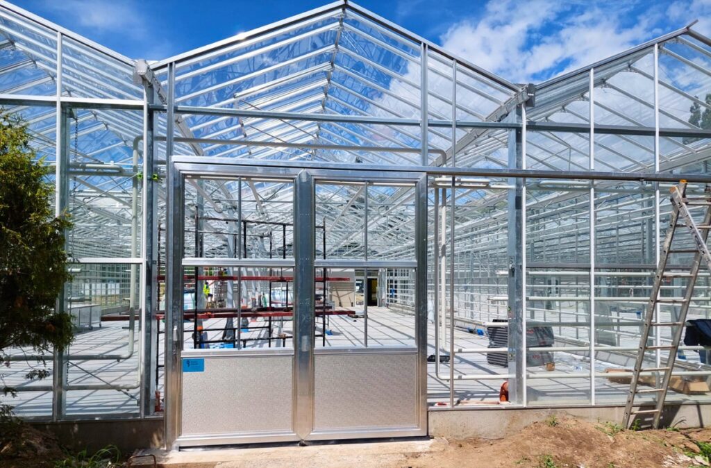 Wejście do szklarni doświadczalnej z pełnym przeszkleniem i konstrukcją stalową.