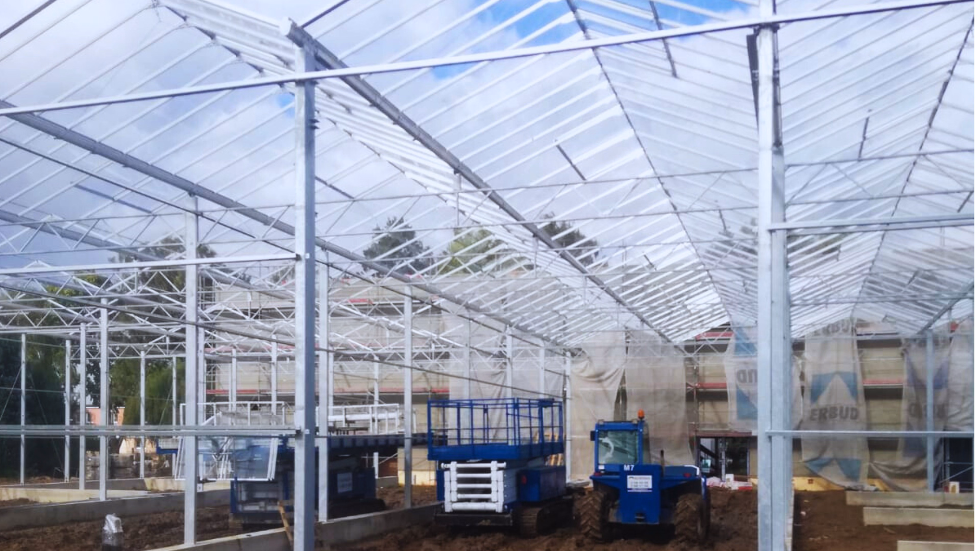 Wnętrze szklarni w Lublinie w trakcie budowy – stalowa konstrukcja nośna, kratownice dachowe i przeszklony dach, widoczne maszyny montażowe.