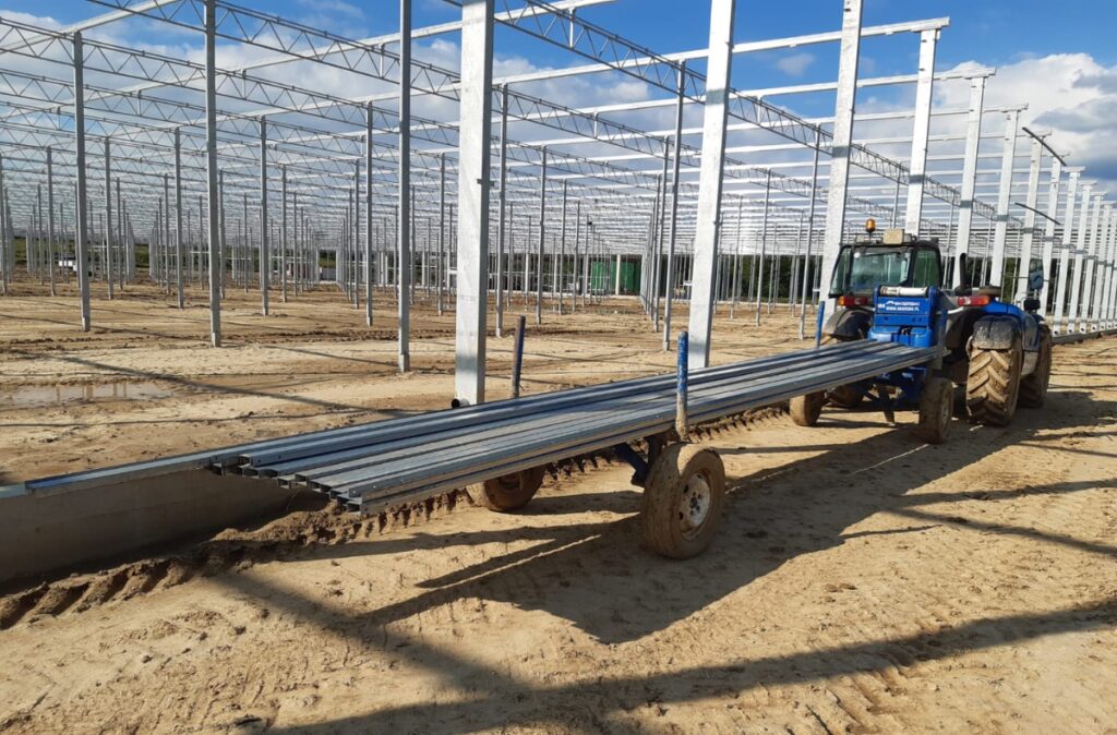 Ciągnik rolniczy z przyczepą transportującą profile stalowe na placu budowy szklarni.