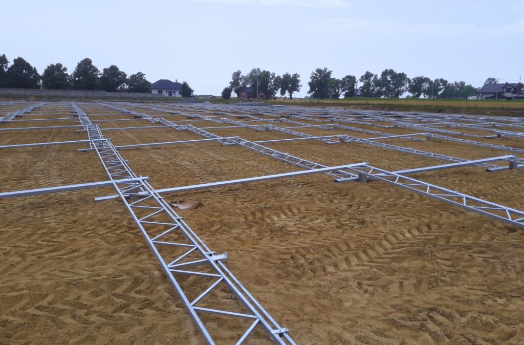 Rozłożone na placu budowy moduły konstrukcji szklarni – stalowe belki i kratownice przygotowane do montażu na wcześniej wyrównanym terenie.